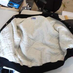 May include: Black and white varsity jacket with a quilted white interior. The jacket has a black collar, cuffs, and hem. The interior is a diamond-patterned quilted material. The jacket is open, displaying the interior.