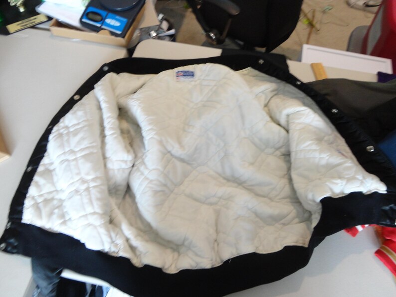 May include: Black and white varsity jacket with a quilted white interior. The jacket has a black collar, cuffs, and hem. The interior is a diamond-patterned quilted material. The jacket is open, displaying the interior.