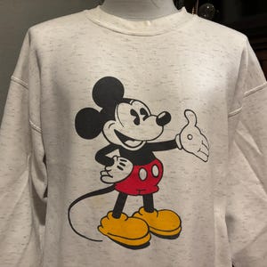May include: A light gray sweatshirt featuring a large graphic of Mickey Mouse. Mickey is depicted in his classic pose, with black ears, a black body, red shorts, yellow shoes, and white gloves. The sweatshirt has a speckled pattern.