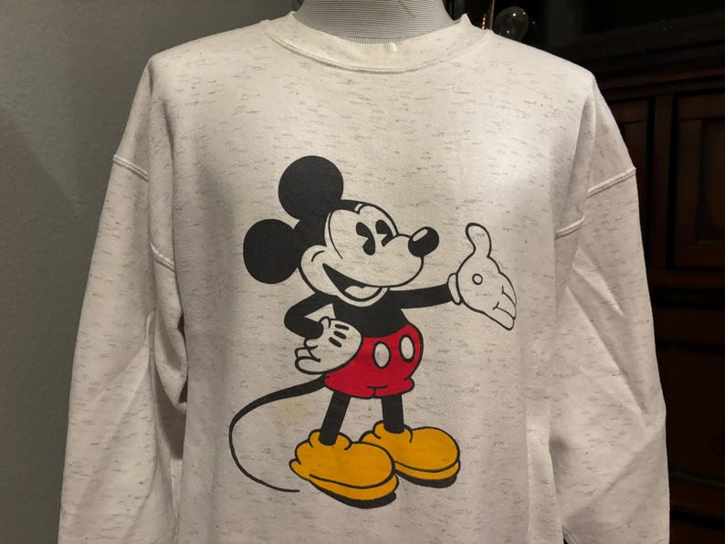 May include: A light gray sweatshirt featuring a large graphic of Mickey Mouse. Mickey is depicted in his classic pose, with black ears, a black body, red shorts, yellow shoes, and white gloves. The sweatshirt has a speckled pattern.