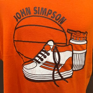 May include: Orange t-shirt with a basketball, a sneaker, and a water bottle printed on it. The text "JOHN SIMPSON" is printed above the basketball.