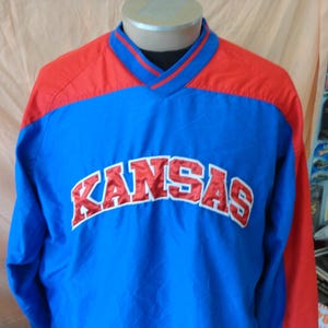 May include: A blue and red pullover jacket with the word "KANSAS" in white and red lettering across the chest. The jacket has a V-neck collar and long sleeves. The jacket is displayed on a mannequin.