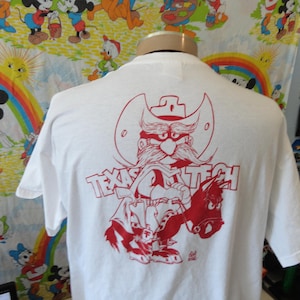 Peut inclure: T-shirt blanc avec un dessin rouge d'un cowboy à grosse moustache et l'inscription "Texas Tech". Le cowboy est à cheval. Le t-shirt est sur un mannequin.