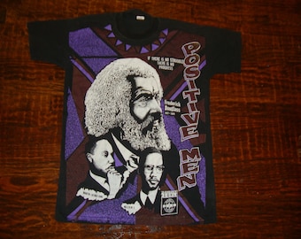 Pode incluir: Camiseta preta com um gráfico de Frederick Douglass e outras figuras históricas. O design inclui o texto "POSITIVE MEN" e "IF THERE IS NO STRUGGLE THERE IS NO PROGRESS". A camiseta tem gola redonda.