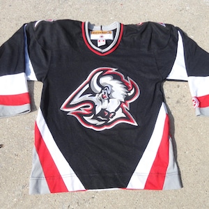 May include: Black hockey jersey with a team logo on the front. The jersey has red, white, and gray accents on the sleeves and collar. The team logo is a white bison with red and gray details.