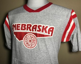 Peut inclure: T-shirt à manches courtes grise avec des manches rayées rouge et blanc. Le t-shirt a un col en V rouge et un graphique rouge et blanc qui dit "NEBRASKA" avec un sceau rouge et blanc qui dit "Universitas Nebraskensis, Feb. 15, 1869".