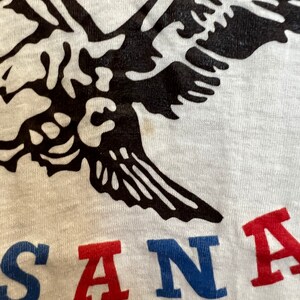 K&ouml;nnte beinhalten: Ein wei&szlig;es T-Shirt mit einem schwarzen Adler-Motiv und dem Wort "SANTA" in Rot, Blau und Wei&szlig;.