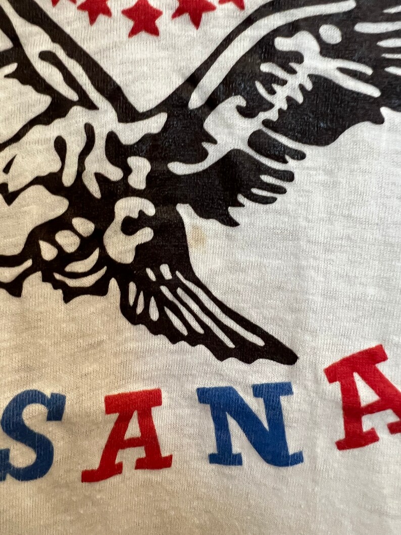 K&ouml;nnte beinhalten: Ein wei&szlig;es T-Shirt mit einem schwarzen Adler-Motiv und dem Wort "SANTA" in Rot, Blau und Wei&szlig;.