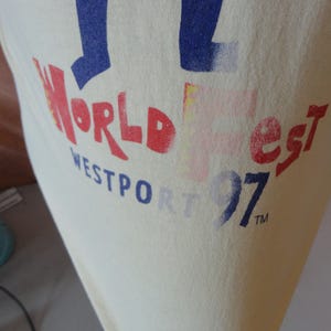 May include: Cream-coloured t-shirt featuring the text "World Fest Westport 97" in red and blue. The print has a slightly faded, vintage appearance. The design is positioned on the front of the shirt.