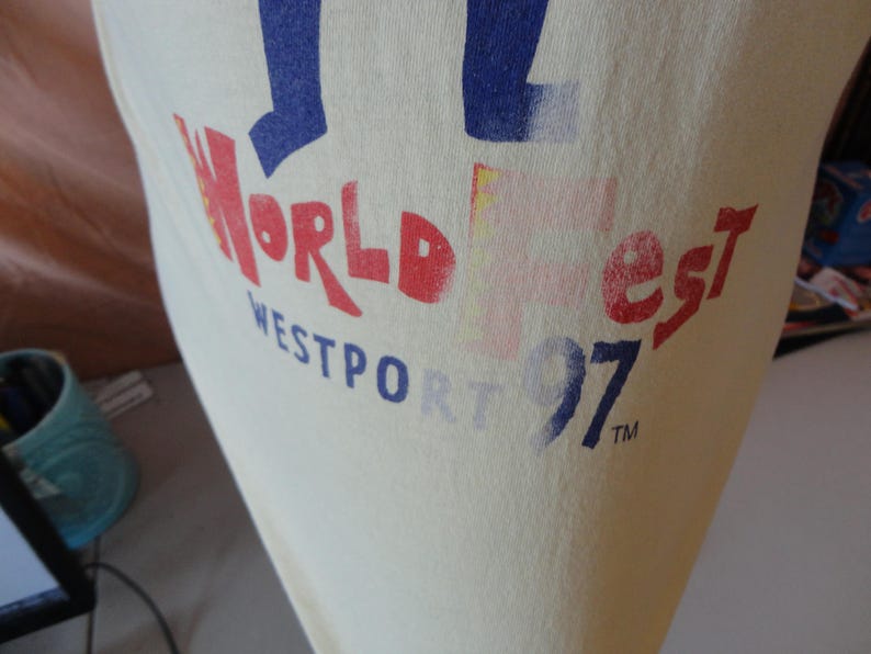 May include: Cream-coloured t-shirt featuring the text "World Fest Westport 97" in red and blue. The print has a slightly faded, vintage appearance. The design is positioned on the front of the shirt.