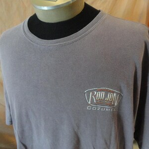 May include: A gray t-shirt with the embroidered text "Ron Jon Surf Shop Cozumel" on the front. The shirt has short sleeves and a crew neck. The shirt is displayed on a mannequin.
