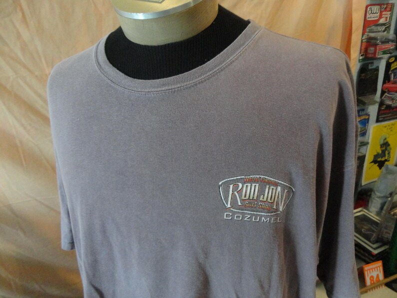 May include: A gray t-shirt with the embroidered text "Ron Jon Surf Shop Cozumel" on the front. The shirt has short sleeves and a crew neck. The shirt is displayed on a mannequin.