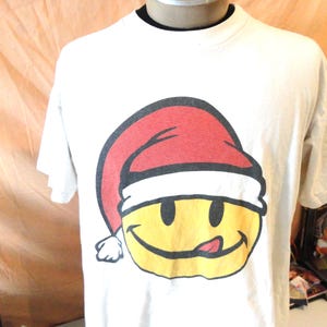 Puede incluir: Camiseta blanca con una carita sonriente amarilla que lleva un gorro de Papá Noel rojo y blanco. La carita sonriente tiene un contorno negro, ojos negros y una lengua rosa. La camiseta tiene un cuello negro.
