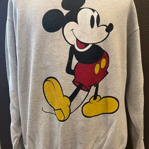 May include: A light gray sweatshirt featuring a large graphic of Mickey Mouse. Mickey is depicted in his classic pose, with black ears, a white face, red shorts, yellow shoes, and black arms. The sweatshirt has a crew neck and long sleeves.