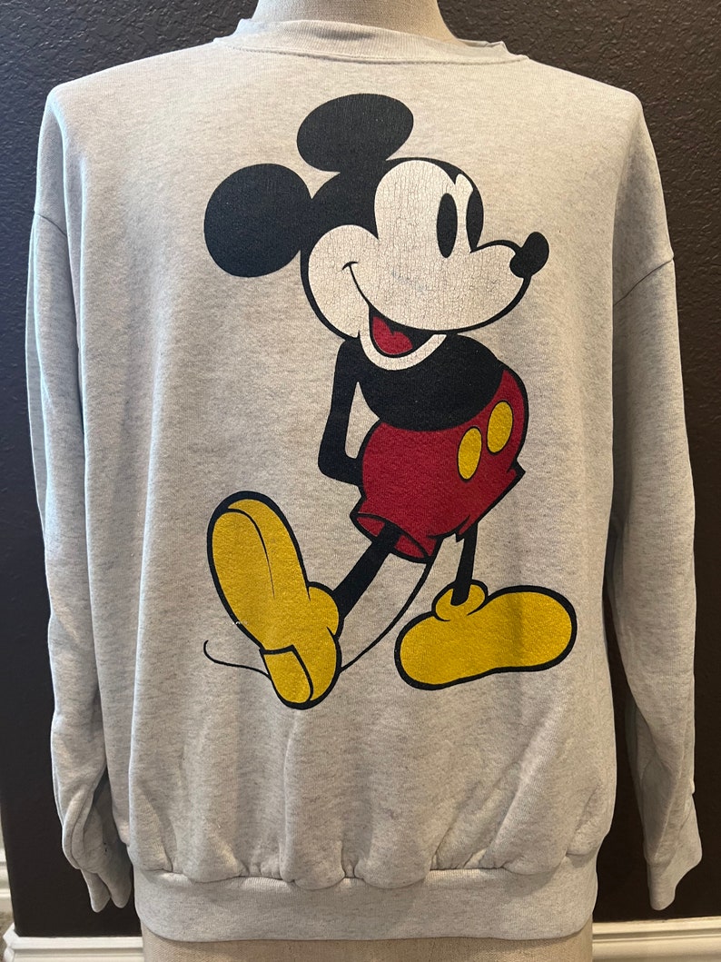 May include: A light gray sweatshirt featuring a large graphic of Mickey Mouse. Mickey is depicted in his classic pose, with black ears, a white face, red shorts, yellow shoes, and black arms. The sweatshirt has a crew neck and long sleeves.