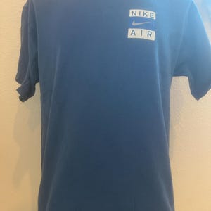 Puede incluir: Una camiseta azul con un logotipo blanco de Nike Air en la parte delantera.