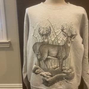 Puede incluir: Una sudadera gris con un estampado gráfico de dos ciervos en un bosque nevado. Los ciervos son marrones y tienen cuernos.