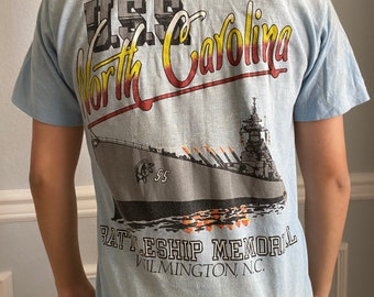 Puede incluir: Una camiseta azul claro con un gráfico del USS North Carolina Battleship Memorial en Wilmington, Carolina del Norte. El gráfico presenta un acorazado gris con llamas rojas, amarillas y naranjas.