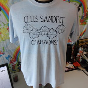 May include: Light blue t-shirt with the text "ELLIS SANDPIT CHAMPIONS!" in black. The shirt features a black floral design above the word "CHAMPIONS!". The shirt is displayed on a mannequin.