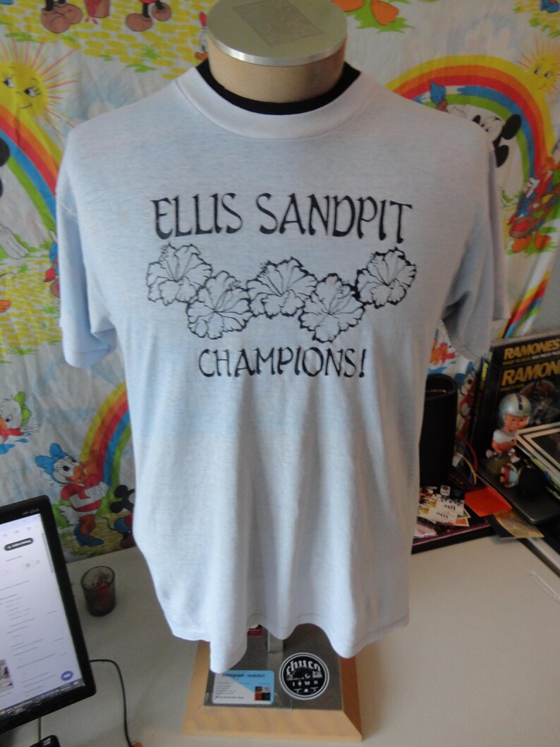 May include: Light blue t-shirt with the text "ELLIS SANDPIT CHAMPIONS!" in black. The shirt features a black floral design above the word "CHAMPIONS!". The shirt is displayed on a mannequin.