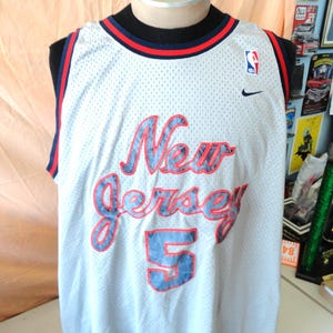 May include: White basketball jersey with the words "New Jersey" in red and blue script, and the number "5" in blue. Features the NBA and Nike logos. The jersey has a black and red trim.