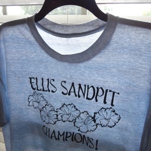 May include: Light blue t-shirt with a gray collar and the text "ELLIS SANDPIT CHAMPIONS!" in black script. Below the text are five black floral designs. The shirt is hanging on a black hanger.