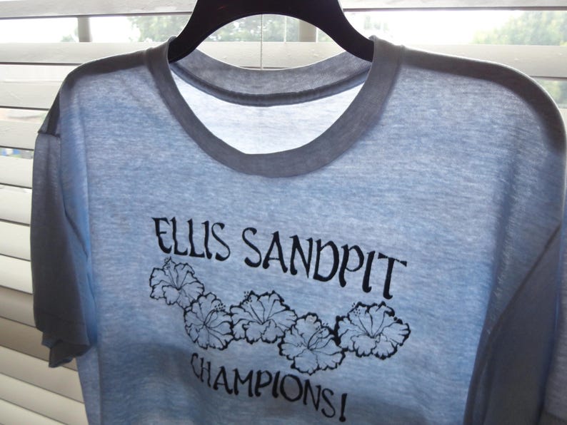May include: Light blue t-shirt with a gray collar and the text "ELLIS SANDPIT CHAMPIONS!" in black script. Below the text are five black floral designs. The shirt is hanging on a black hanger.