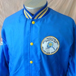 以下が含まれることがあります： 黄色と白のストライプの襟と黄色のボタンが付いた青いナイロンジャケット。「ARCO PRODUCTION DEPT PRUDHOE BAY」と「MILLION SAFE MANHOURS」の文字が入った丸いパッチが特徴です。左袖にはアメリカ国旗のパッチが見えます。