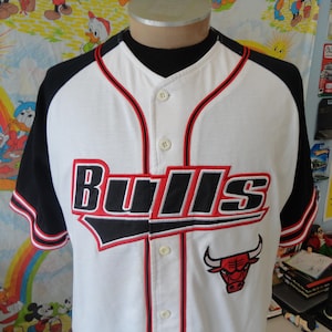 Op de afbeelding: Wit baseballshirt met zwarte en rode bies. Het shirt heeft het woord "Bulls" in zwarte en rode letters met een rood stierenlogo op de voorkant.
