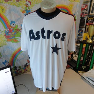 Puede incluir: Camiseta de béisbol blanca con la palabra "Astros" en azul marino en el pecho, junto con una estrella azul marino. La camiseta tiene cuello en V y ribetes azul marino y rojo. La camiseta se exhibe en un maniquí.