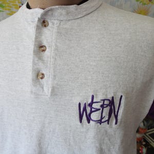 May include: A light gray Henley shirt with purple sleeves. The shirt has three brown buttons and the embroidered letters "WEPN" in purple on the chest. The shirt is made of a soft, comfortable material.