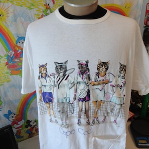Op de afbeelding: Wit T-shirt met een afbeelding van antropomorfe katten in tennisoutfit. De katten zijn afgebeeld in verschillende poses, met de tekst "Cape Cod" eronder. Het shirt heeft een ronde hals en korte mouwen.
