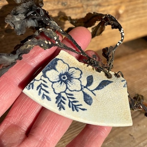 May include: A broken piece of white ceramic with a blue floral pattern. The fragment is held in a hand, with dried seaweed. The background is a wooden surface.