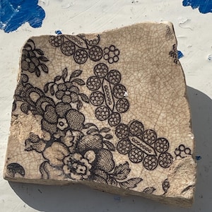 May include: A fragment of antique ceramic tile with a floral pattern in black and cream. The design features stylized flowers and circular motifs, with visible cracks across the surface. The tile's edges are irregular, suggesting it's a broken piece.