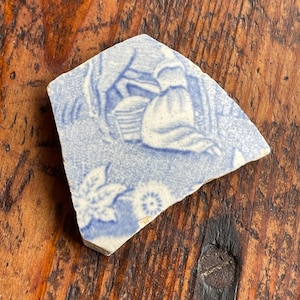 May include: A fragment of antique blue and white ceramic with a pastoral scene. The design features a seated figure and foliage. The broken edge reveals the ceramic's thickness. The piece rests on a wooden surface.