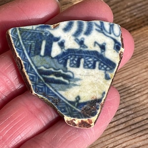 May include: A fragment of antique blue and white ceramic pottery. The design depicts a bridge and figures, with a detailed border. The edges show signs of age and wear, with a brown, aged appearance. The pottery shard is held in a hand.
