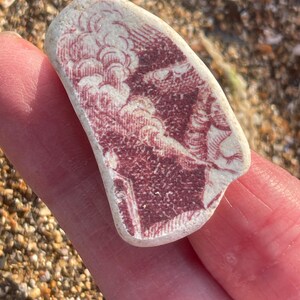 May include: A fragment of sea pottery featuring a faded red and white landscape design. The design includes clouds and buildings. The pottery has a smooth, worn texture, indicating it has been smoothed by the sea. A unique piece of beachcombed art.