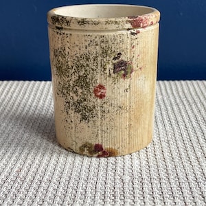 Op de afbeelding: Een cilindrische, gebroken witte steengoed pot met verticale ribbels. De pot heeft een licht getextureerd oppervlak met groene en kastanjebruine bloemaccenten. De rand is een donkerdere tint gebroken wit. De pot staat op een lichtgrijs geweven oppervlak.
