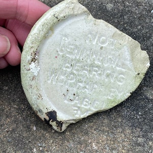 Può includere: Frammento di oggetto in ceramica bianca con la scritta in rilievo "NOT GENUINE BEARING W.P.A.R.C. LABEL FM". L'oggetto ha una forma arrotondata con un bordo rotto e segni di usura.