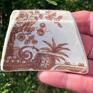 May include: A fragment of antique ceramic with a brown transferware design. The design features floral and botanical elements, including a tree, vines, and a pineapple. The piece has a white background and is held in a hand.