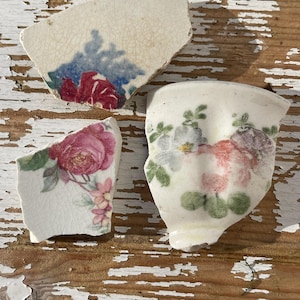 May include: Three pieces of broken white ceramic with floral patterns. The pieces are on a whitewashed wooden surface. The floral patterns are in shades of pink, red, blue, and green.