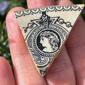 Puede incluir: Una pieza triangular de azulejo de cerámica con un diseño en blanco y negro. El diseño presenta el perfil de una mujer en un círculo rodeado de detalles ornamentados.