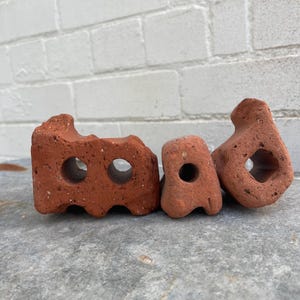 Puede incluir: Tres ladrillos de arcilla roja con agujeros. Los ladrillos están dispuestos en una fila de izquierda a derecha. Los ladrillos están sobre una superficie gris.