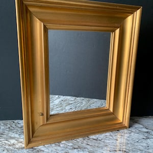May include: A gold-colored rectangular picture frame with a beveled edge. The frame is made of wood and has a glossy finish. The frame is sitting on a marble surface.