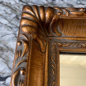 May include: A rectangular mirror featuring a thick, ornate frame with a metallic gold finish. The frame displays intricate, raised scrollwork and carved detailing along the edges, with a smooth, reflective glass surface visible in the center.