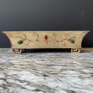 May include: A rectangular brass planter with a decorative floral design. The planter features a copper-colored vine with green and red jewel-toned accents. The planter is supported by four small, square feet.