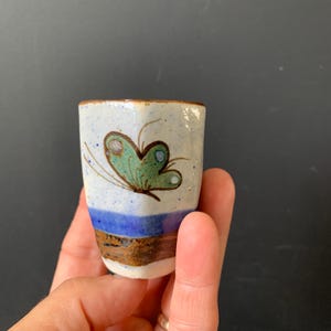 May include: A small ceramic cup with a speckled off-white glaze featuring a hand-painted green butterfly design. The base of the cup is decorated with horizontal bands of solid blue and brown glaze, creating a distinct contrast against the speckled body.