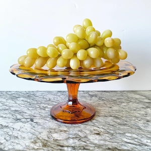 May include: An amber-colored glass cake stand with a scalloped edge, holding a bunch of green grapes. The stand has a pedestal base with a star-shaped design. The grapes are ripe and ready to eat.