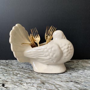 May include: A cream-colored ceramic dove-shaped utensil holder with a fan-shaped tail. The dove holds several gold-colored forks. The holder is on a marble-patterned surface with a dark background.