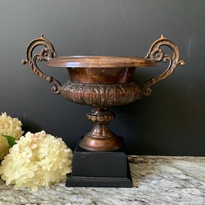 May include: A metallic copper-toned urn with two ornate, scrolled handles and a wide, flared rim. The body features a decorative leaf-patterned band, supported by a pedestal base resting on a square, solid black block stand.